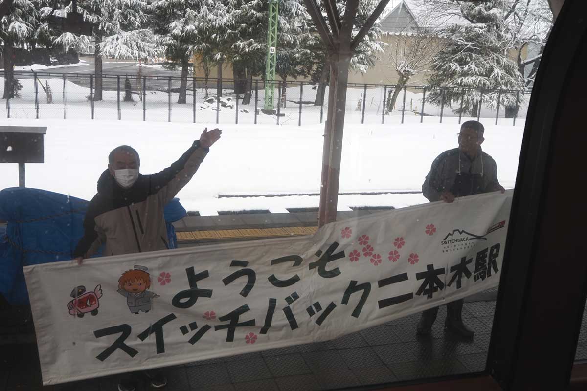 二本木駅で「ようこそ　スイッチバック二本木駅」の横断幕を持ち、雪月花を出迎える地元住民（大塚圭一郎撮影）